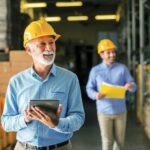 Picture_of_father_and_son_walking_through_their_warehouse_with_helmets_on_their_heads._Father_is_holding_digital_tablet_in_his_hand_and_looking_at_shelf_full_of_boxes_with_smile_on_his_face.